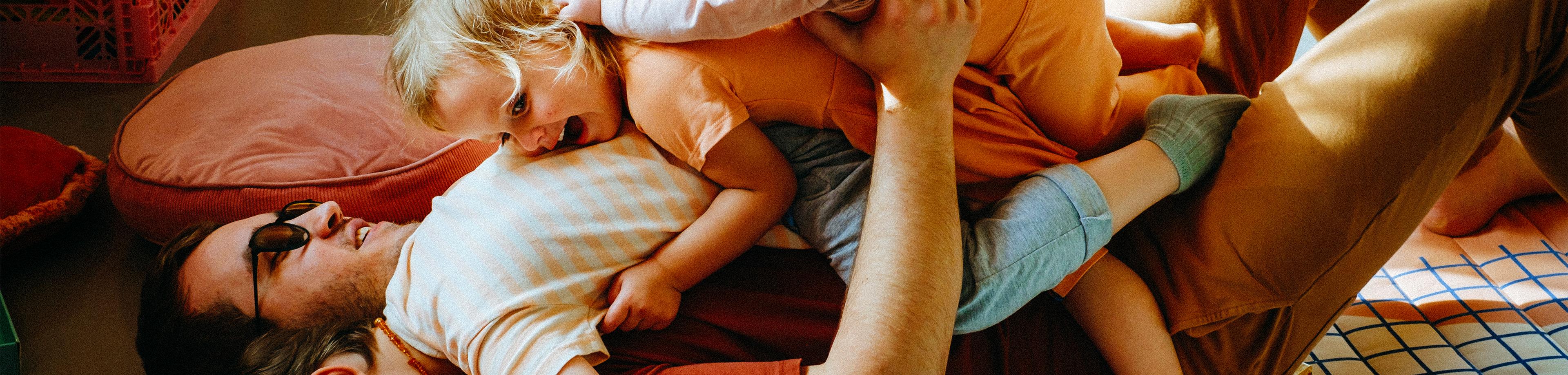 Three young children playfully lay on top of their father on the floor at home