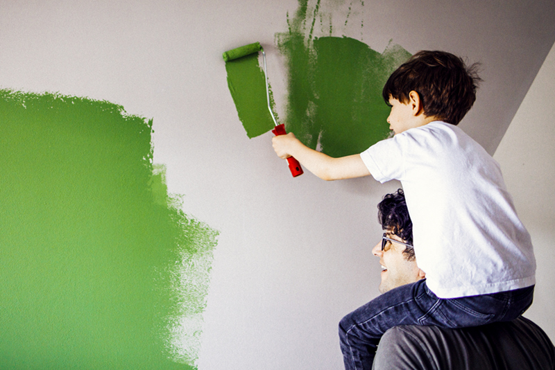 A young boy sits on a man his shoulders and uses a paint roller to paint the wall green