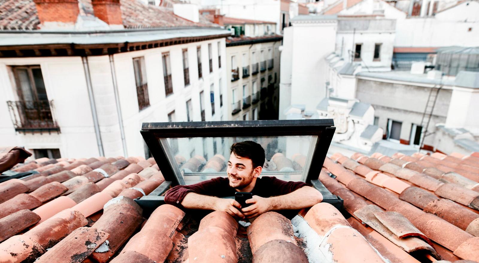 Large Web and Screen A happy young man sticks his head out of skylight to look at the city view with his phone in his hands