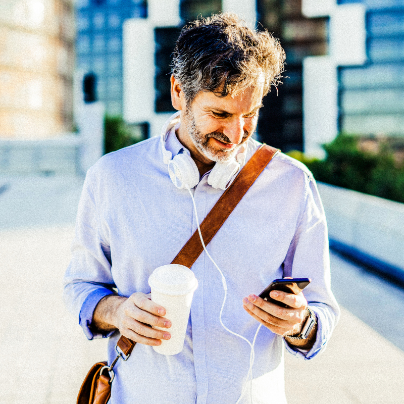 Mid adult man holding coffee cup looking at mobile phone with headphones around neck