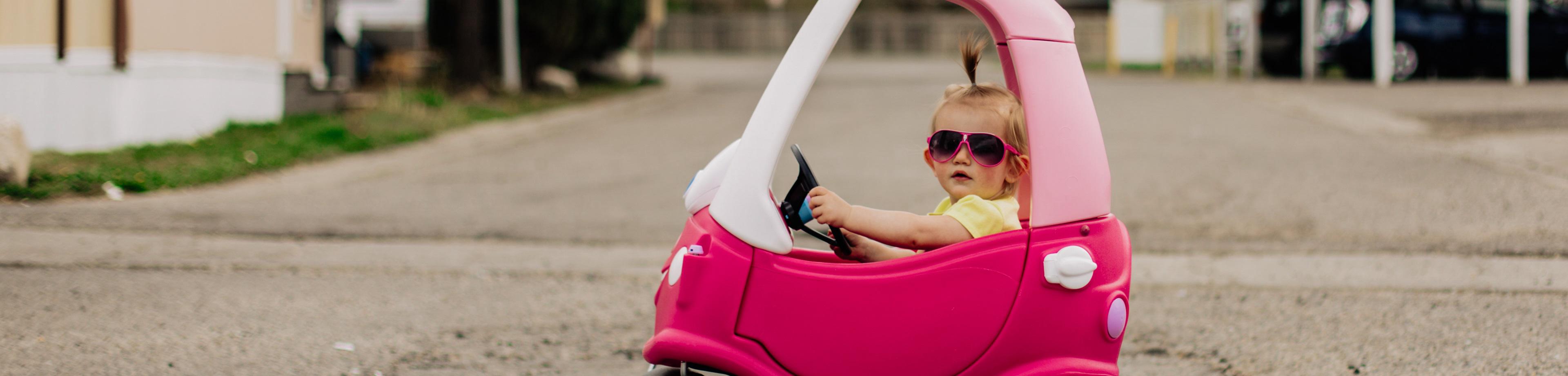 Young girl in sunglasses driving a plastic toy car outside